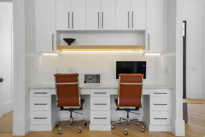 Dual-desk home office featuring built-in white cabinetry with ample storage drawers, integrated task lighting with warm-toned LED strips, and a spacious work surface