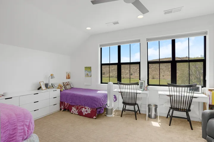 Vaulted ceiling room featuring two large windows with black grilles, light beige carpet, and a ceiling fan