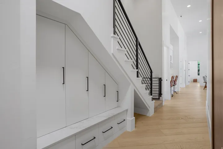 Modern hallway featuring custom white built-in cabinetry with integrated drawers and hardware accents, engineered wood-finish flooring throughout, recessed ceiling lighting, and a contemporary black metal stair railing