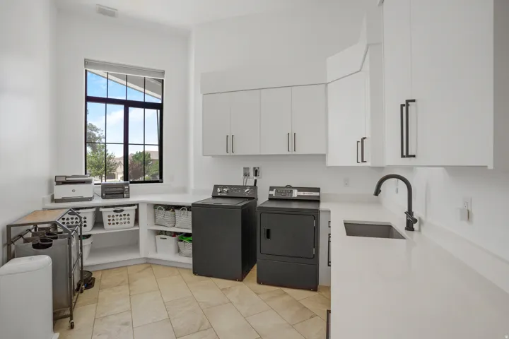 Dedicated laundry room featuring stacked washer and dryer units, white cabinetry with ample storage, tile flooring, a utility sink with modern faucet, and a large multi-pane window providing natural light and outdoor views
