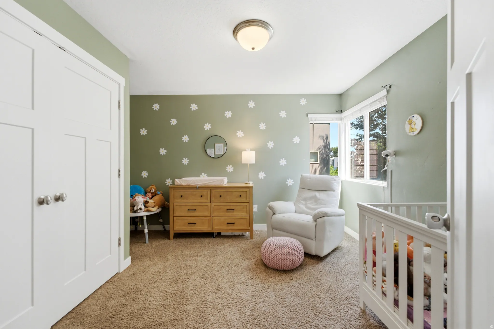 Versatile nursery featuring soft sage-green walls adorned with decorative daisy motifs, natural wood-finish dresser with multiple drawers, white crib with railings, cushioned seating chair, cork-finish flooring, recessed ceiling lighting, and ample closet