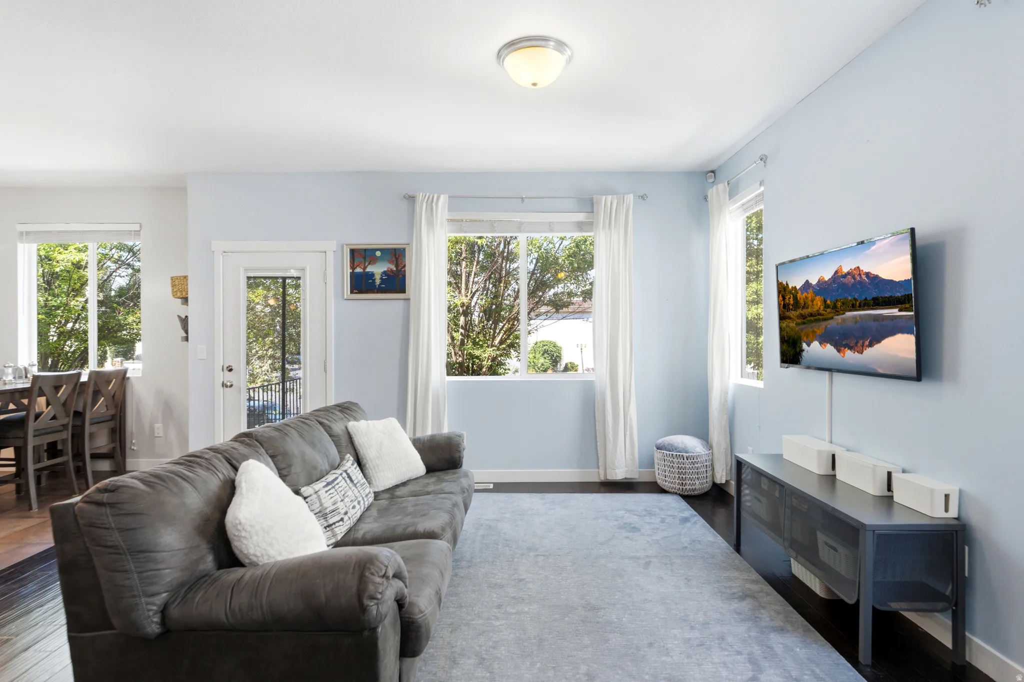 Bright and airy living room featuring light blue walls, expansive windows with natural light, wood-finish flooring, a comfortable gray upholstered sofa with decorative pillows, and a wall-mounted flat-screen television above a dark media console