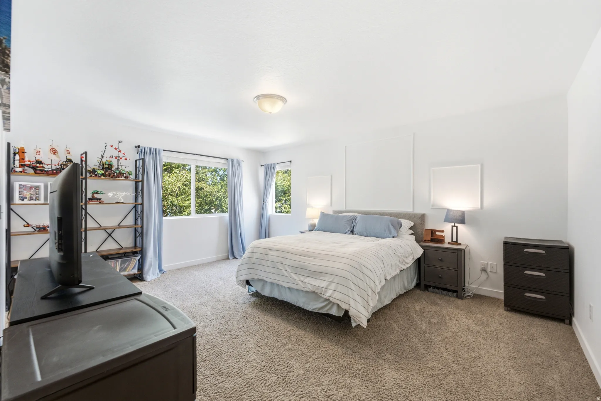 Spacious primary bedroom featuring abundant natural light from dual windows, neutral carpet flooring, and coordinating wood-finish nightstands with integrated task lighting