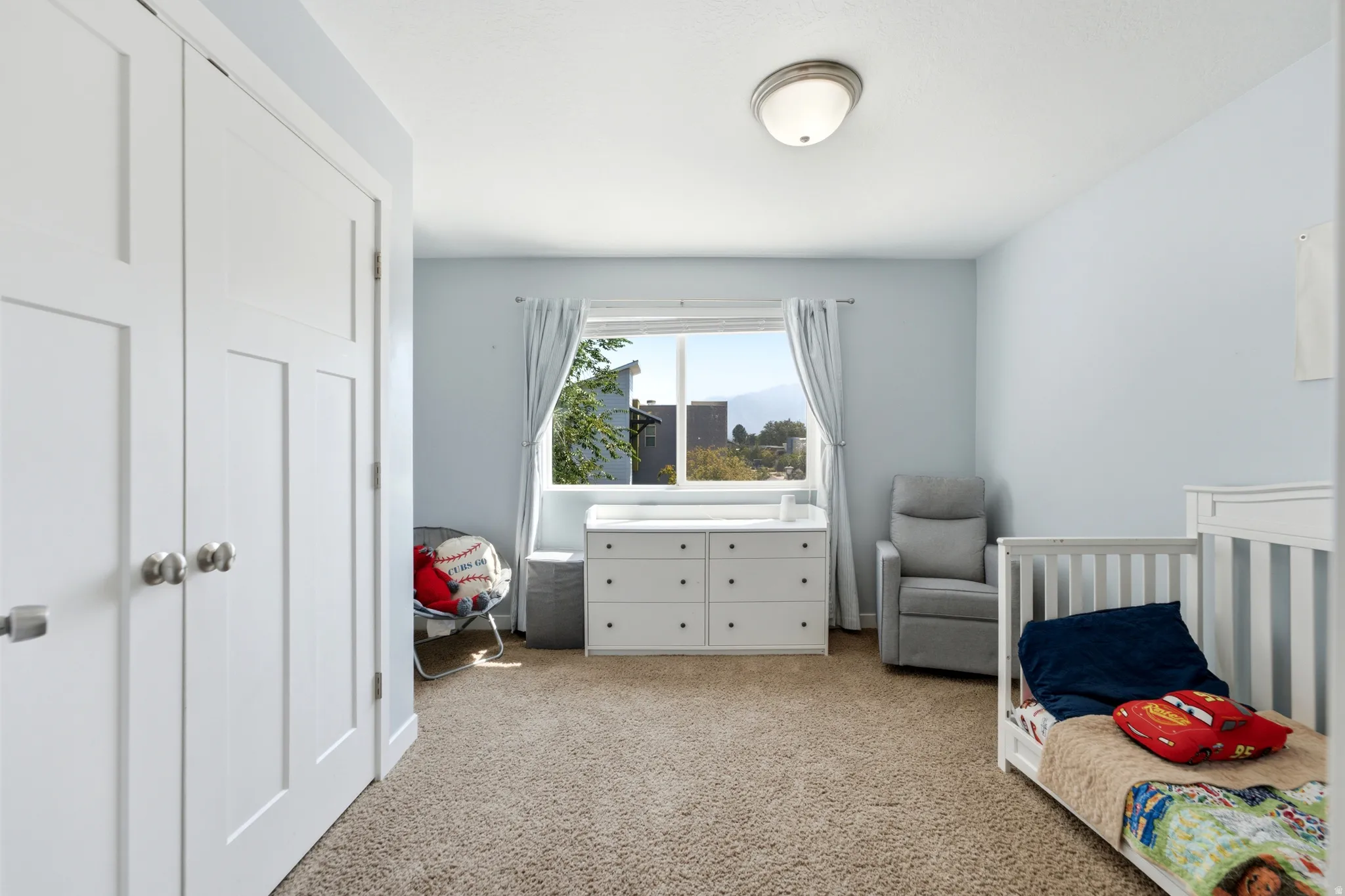 Bright and spacious bedroom featuring carpet flooring, abundant natural light from a large picture window with curtains, white six-drawer dresser with attached top surface, white built-in closet cabinetry, ceiling-mounted light fixture, and soft blue-gray