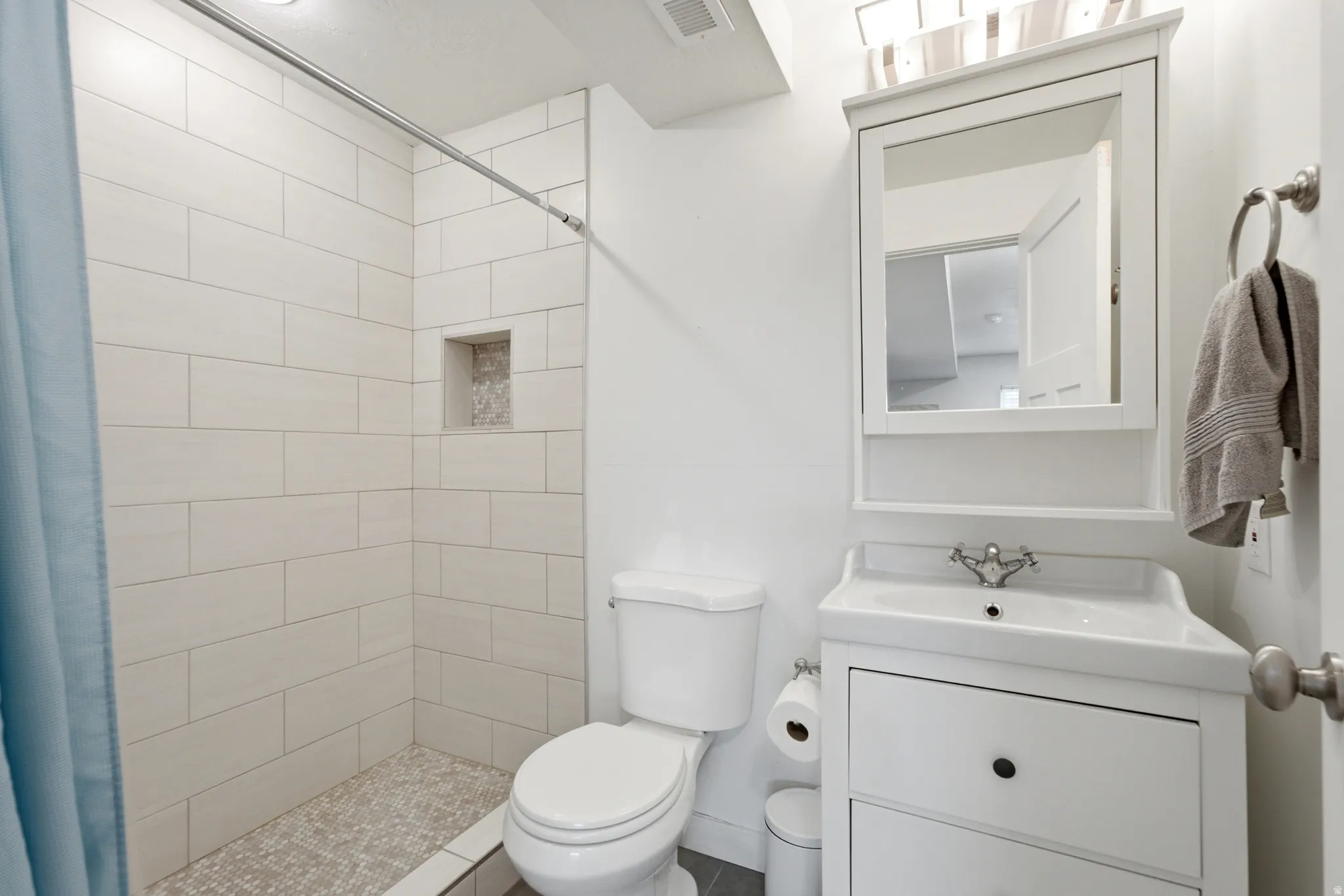 Modern bathroom featuring crisp white subway tile walls, a walk-in shower with frameless glass enclosure and stone-look floor, a white vanity with black hardware and integrated sink, and a toilet with overhead ventilation