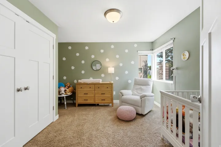 Versatile nursery featuring soft sage-green walls adorned with decorative daisy motifs, natural wood-finish dresser with multiple drawers, white crib with railings, cushioned seating chair, cork-finish flooring, recessed ceiling lighting, and ample closet