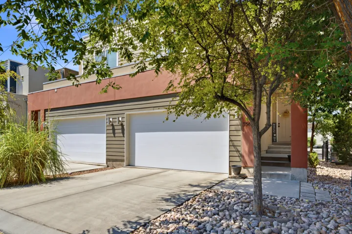 Contemporary brick exterior with dual-panel garage doors, concrete driveway, and mature landscaping featuring ornamental grasses and established trees providing natural shade to the front façade