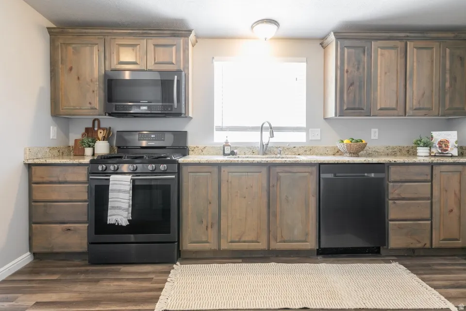 Kitchen featuring wood-finish cabinetry, granite-style countertops, stainless steel sink with gooseneck faucet, black appliances including oven range, microwave, and dishwasher, and wood-finish flooring