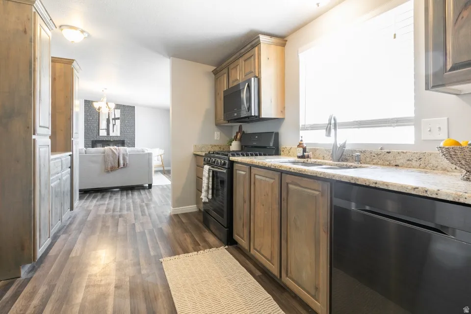 Galley-style kitchen featuring wood-finish cabinetry, granite countertops, stainless steel appliances including a gas range and dishwasher, undermount sink with chrome faucet, and wood-finish flooring that extends throughout the home