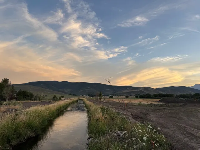 Picturesque rural property featuring an irrigation canal with native grasses and wildflowers lining the banks, expansive open acreage, distant mountain backdrop, and dramatic evening sky with cloud formations