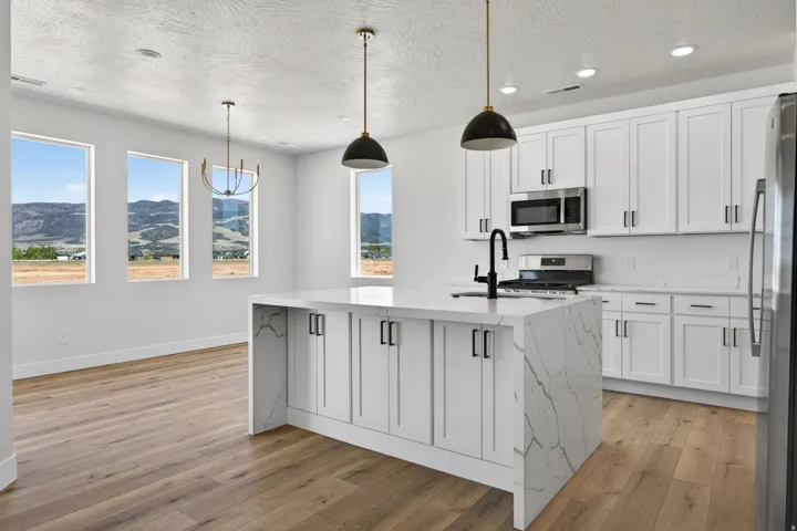 Bright, light-filled kitchen featuring white shaker-style cabinetry, a large waterfall-edge island with marble-look countertops, stainless steel appliances, pendant lighting with matte black fixtures, and wood-finish flooring throughout