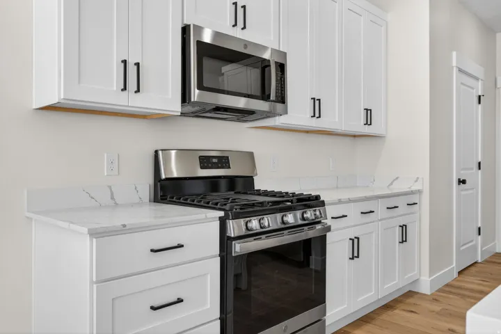 Modern kitchen featuring white shaker-style cabinetry with brushed black hardware, white quartz countertops with subtle veining, stainless steel gas range with six burners, stainless steel over-the-range microwave, and wood-finish flooring throughout