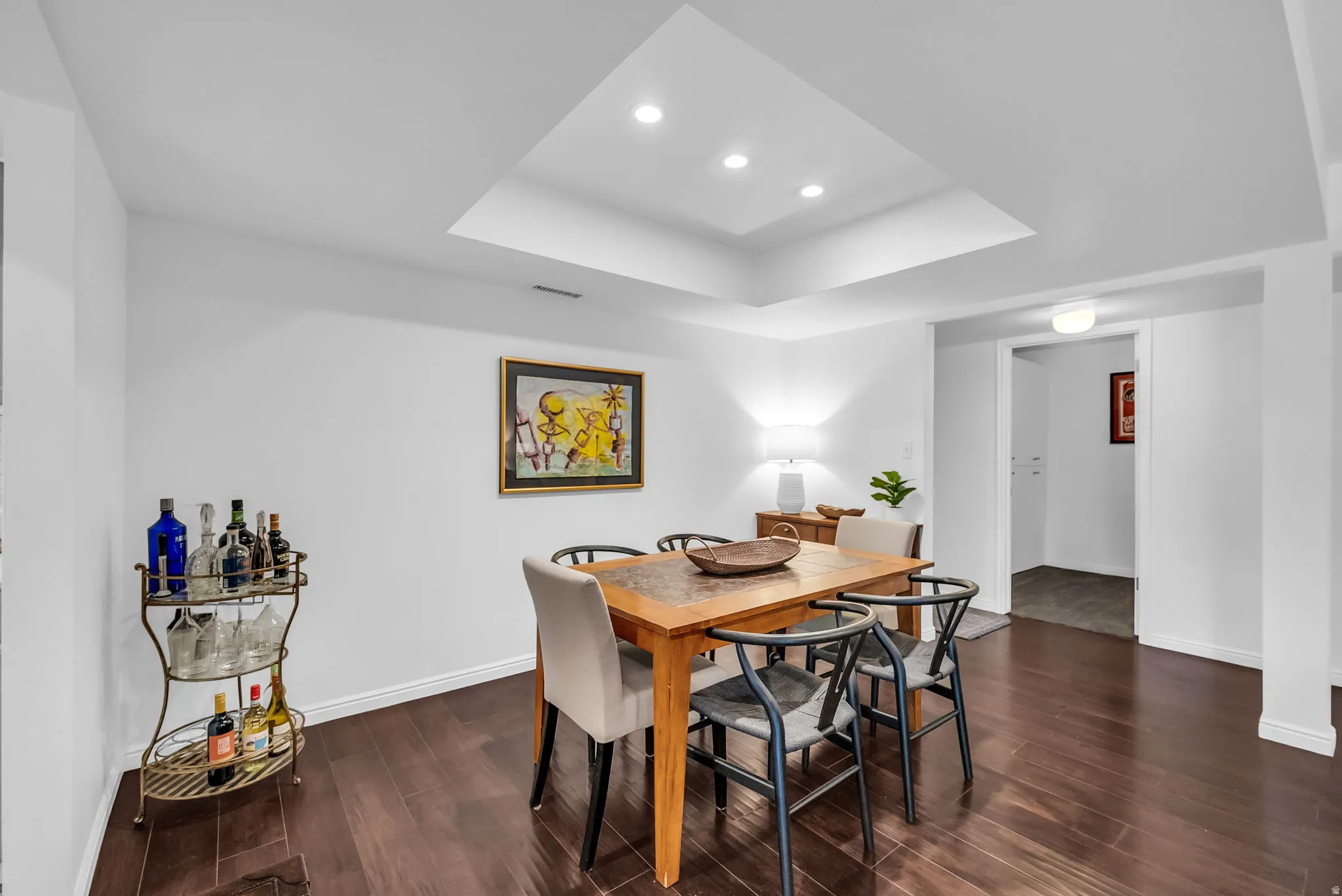 Sophisticated dining area featuring a wood-finish table with coordinating black wishbone chairs, recessed ceiling lighting with a skylight soffit, dark wood-finish flooring, and a beverage cart with metal framework