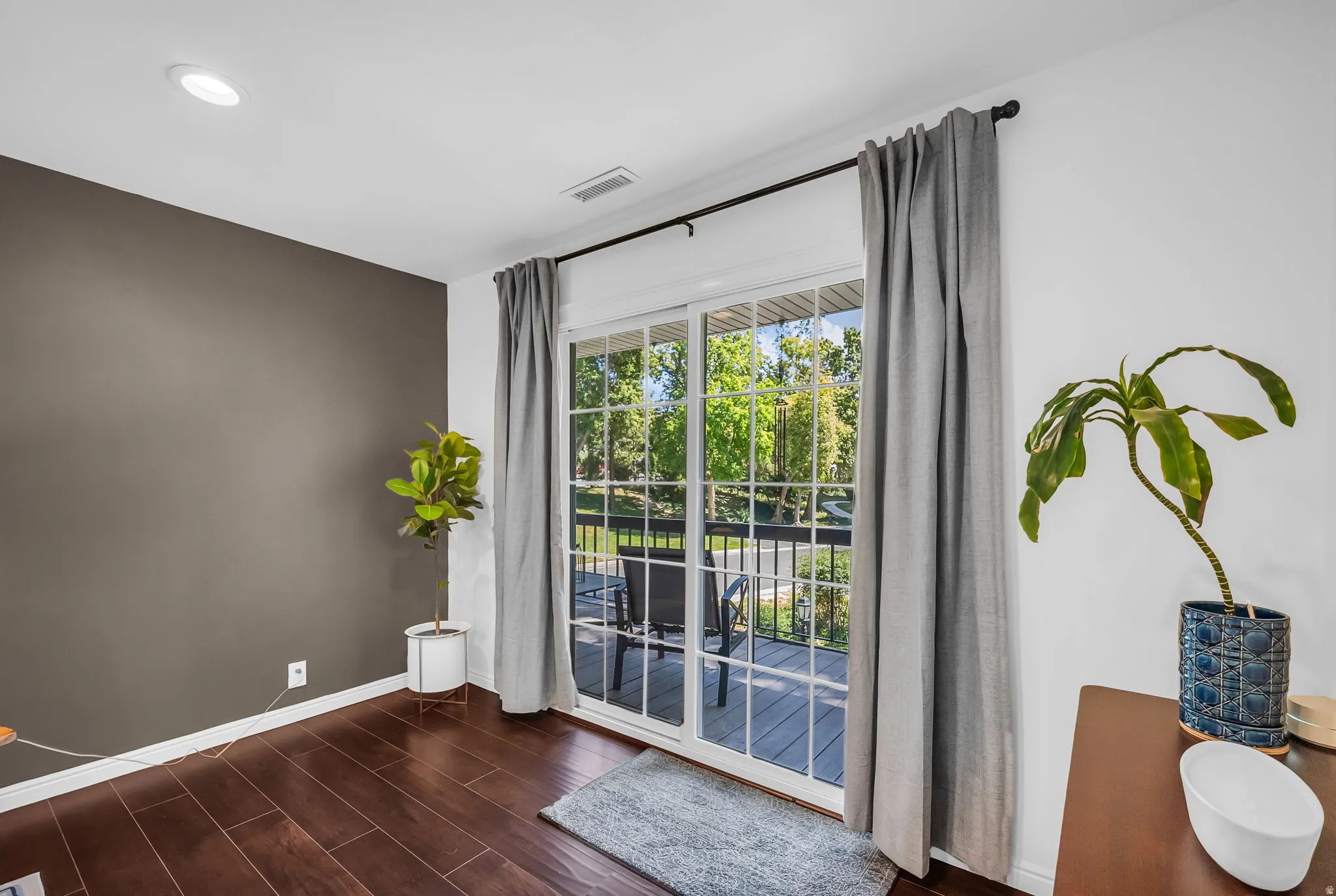 Bright, contemporary room featuring gleaming wood-finish flooring, a dramatic taupe accent wall, and expansive glass sliding doors framed by floor-to-ceiling gray drapery