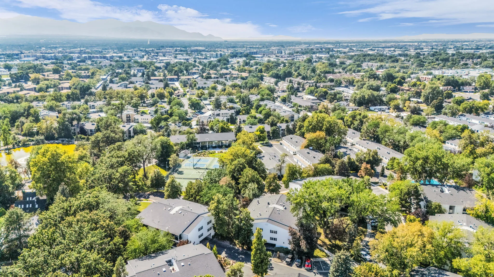 Expansive residential area featuring numerous mature trees, a tennis court, and a body of water, all against a backdrop of distant mountains