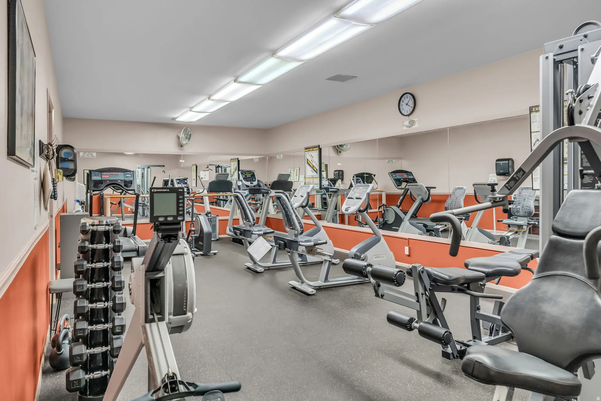 Spacious fitness room featuring a full wall of mirrors, overhead fluorescent lighting, and durable rubber flooring