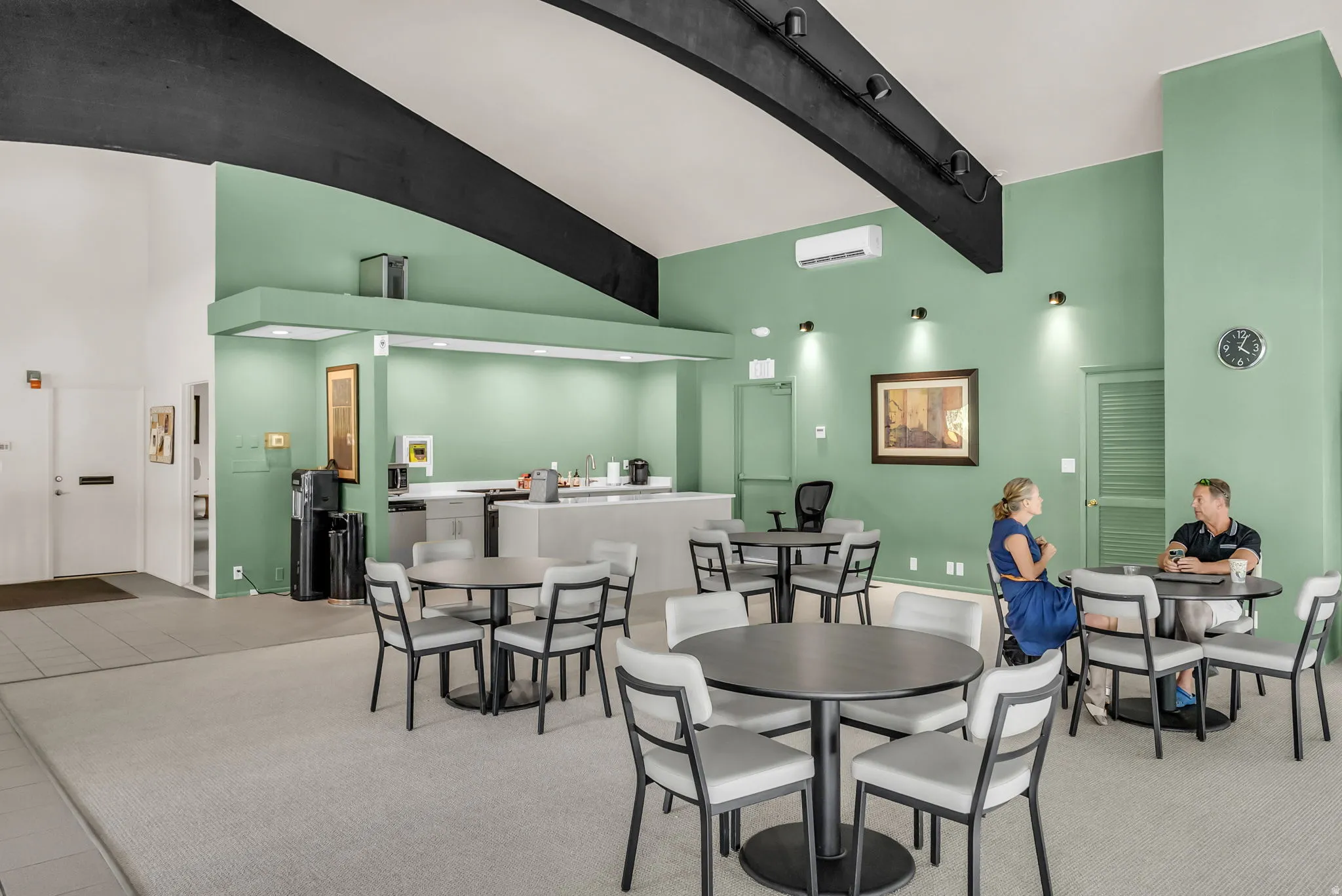 Modern break room and kitchenette featuring sage green accent walls, black exposed beam ceiling detail, and neutral polished concrete flooring
