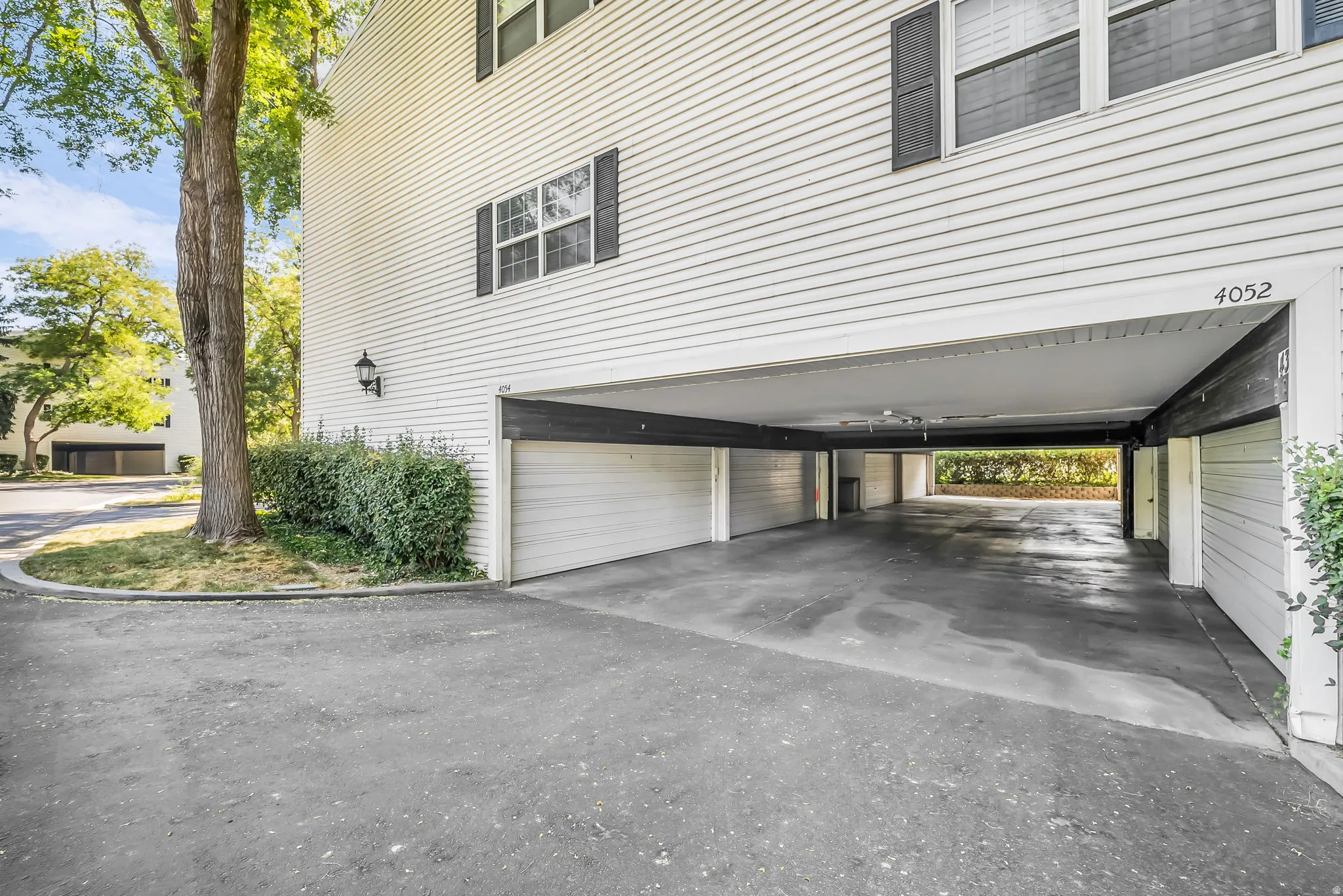 Spacious driveway with extended carport featuring overhead clearance and concrete surface, complemented by mature landscaping and white horizontal siding exterior