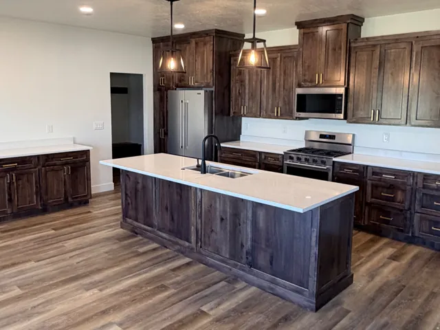 Spacious kitchen featuring a central island with a double basin sink, rich wood-finish cabinetry, light-toned countertops, and wood-finish flooring