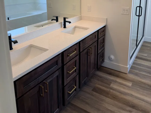 Bathroom vanity featuring dual under-mount sinks with matte black faucets, a spacious quartz countertop, dark wood-finish cabinetry with brass hardware, and wood-finish flooring