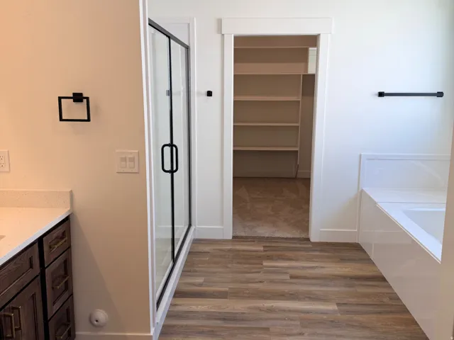 Bathroom featuring wood-finish flooring, a glass-enclosed shower, a built-in bathtub, a vanity with dark wood cabinetry and a light-colored countertop, and a walk-in closet with shelving