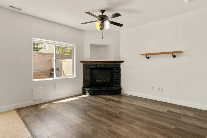 Room featuring wood-finish flooring, a corner fireplace with stone surround and wood mantel, a ceiling fan with integrated lighting, a sliding window, and a wall-mounted wood shelf with pipe brackets