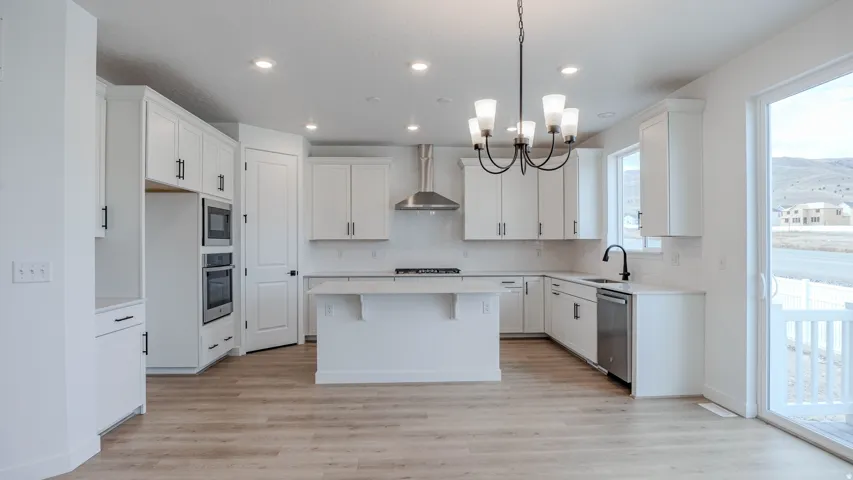 Bright, open-concept kitchen featuring white cabinetry, stainless steel appliances including dual wall ovens, a large center island with seating, wood-finish flooring throughout, recessed and pendant lighting, and sliding glass doors opening to a covered