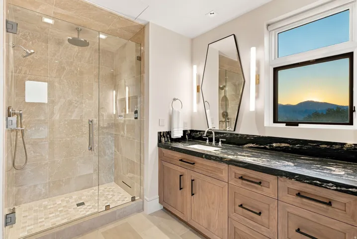 Spacious bathroom featuring a glass-enclosed shower with multiple showerheads, a long vanity with wood cabinetry, a dark stone countertop, a rectangular mirror, and sconce lighting