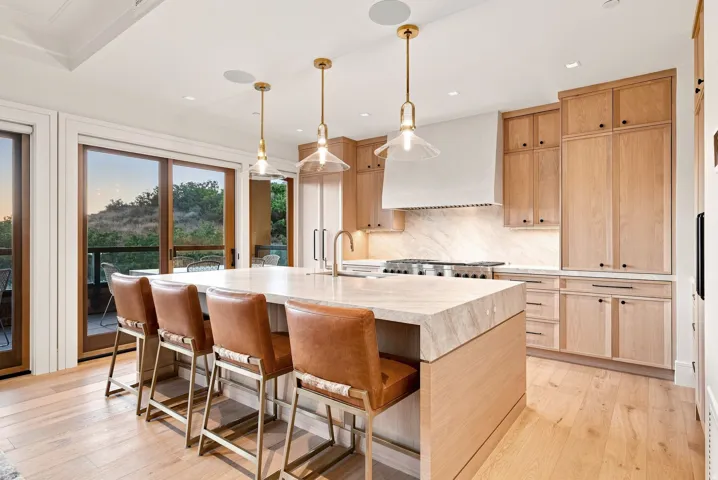 Chef's kitchen featuring natural wood cabinetry with soft-close doors, a large waterfall-edge island with seating for multiple guests, stainless steel gas cooktop, white subway tile backsplash, and quartz countertops