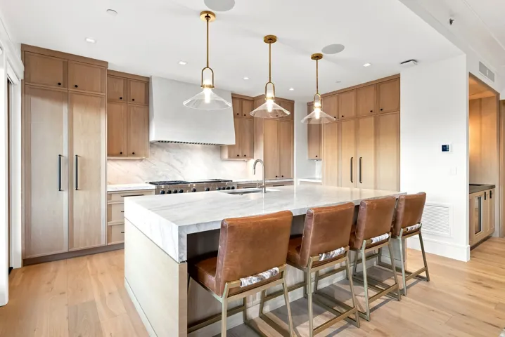 Chef's kitchen featuring custom wood cabinetry, white quartz countertops, a large waterfall-edge island with seating for four, stainless steel gas range with professional-grade hood, pendant lighting with brass fixtures, and wide-plank wood-finish floorin