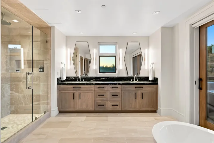 Contemporary bathroom featuring a dual vanity with a dark countertop and two geometric mirrors, a glass-enclosed shower with tiled walls and a rain shower head, large format floor tiling, and a built-in window