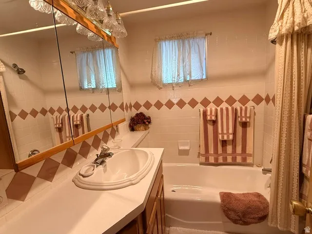 Bathroom featuring a vanity with an integrated sink, a built-in medicine cabinet with integrated lighting, a full bathtub, and tiled walls with a decorative accent band