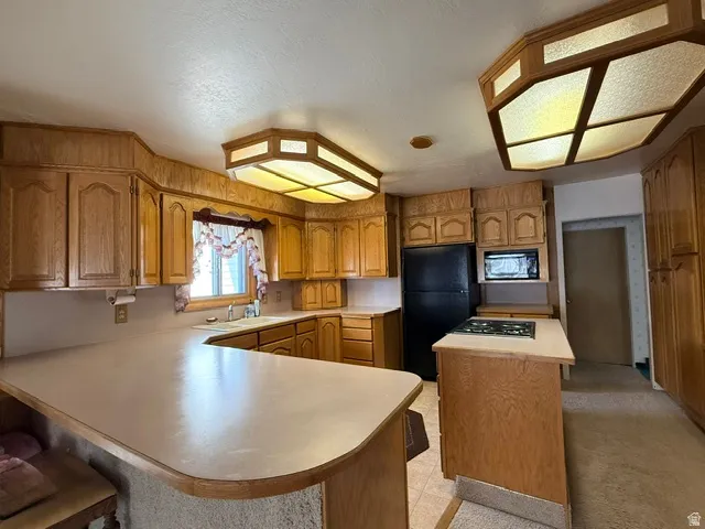 Spacious kitchen featuring warm wood cabinetry throughout, dual ceiling fixtures with geometric frames, stainless steel appliances including a black range and built-in microwave, curved countertops with ample workspace, and a central island with seating
