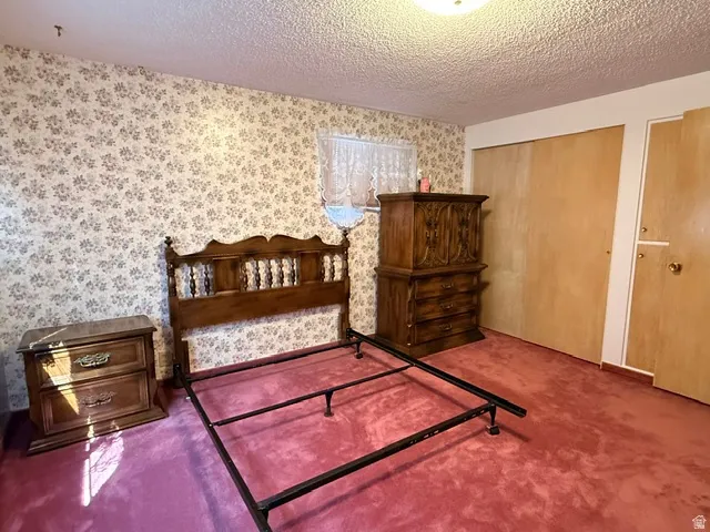 Bedroom featuring floral wallpaper, textured ceiling, a window with sheer curtains, plush carpeting, and built-in closet storage with wood-finish doors