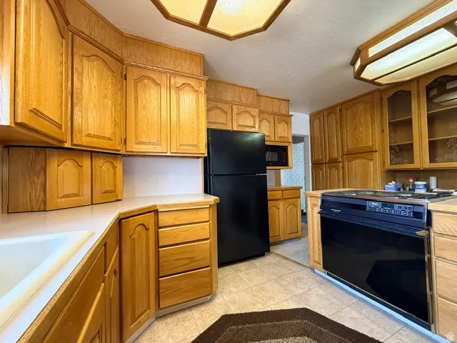 Spacious kitchen featuring warm wood-finish cabinetry throughout, white laminate countertops, black appliances including a side-by-side refrigerator and electric range, recessed ceiling lighting, and tile flooring