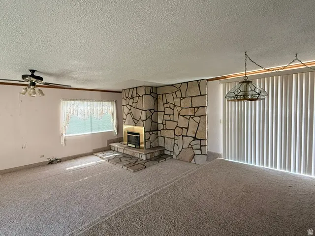Spacious living room showcasing a striking stone fireplace with geometric pattern detailing, carpeted flooring, ceiling fan with light fixture, and dual windows with vertical blinds providing natural light and privacy
