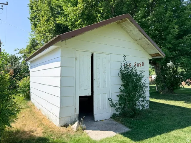 Detached storage shed featuring white horizontal siding, gabled roof with brown trim, double doors with white paneling, and established climbing ivy