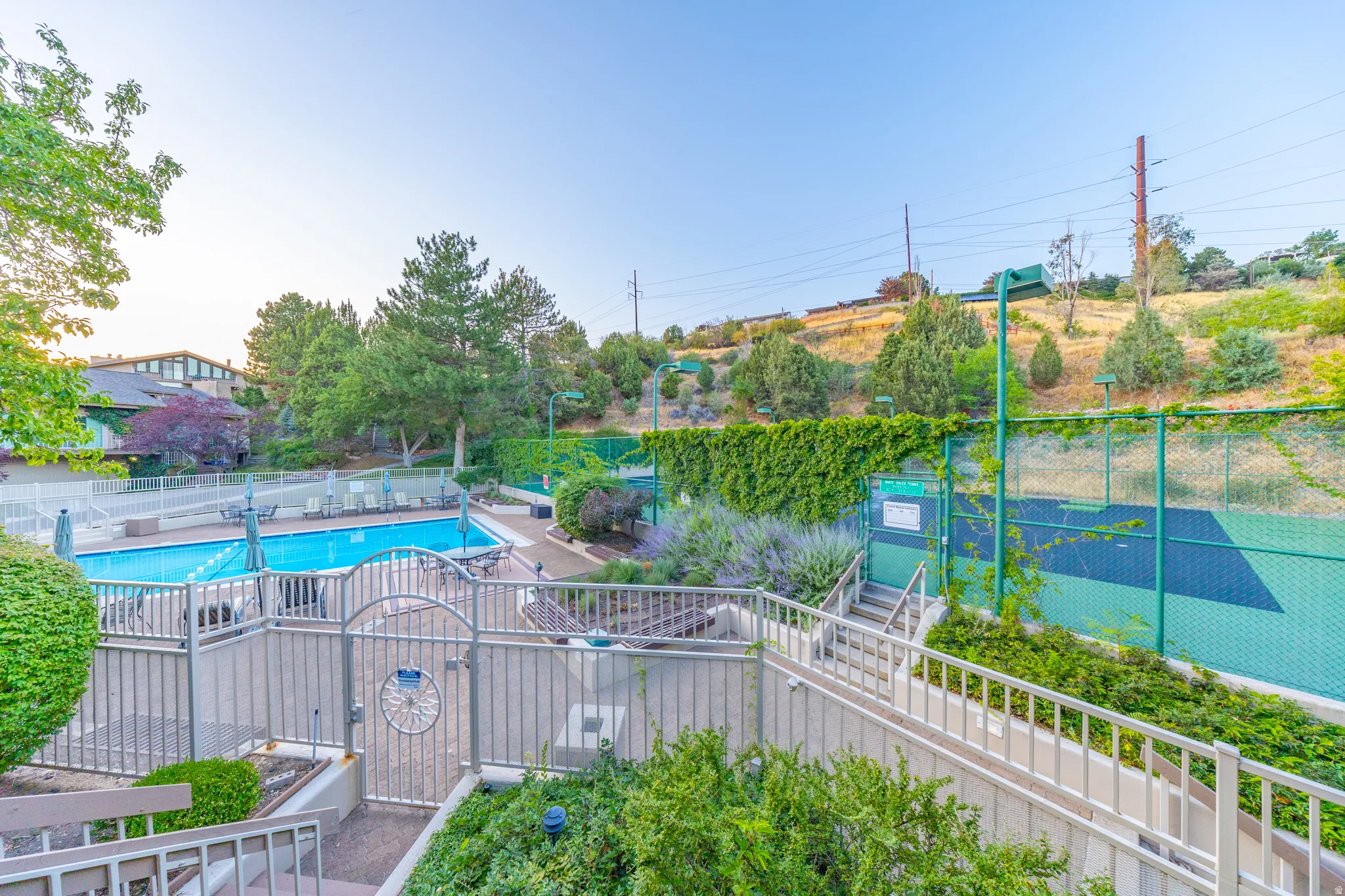 Resort-style outdoor recreation area featuring a sparkling swimming pool with white safety fencing, adjacent tennis court with professional-grade netting, mature landscaping with established trees and shrubs, and expansive hardscaped deck with white raili