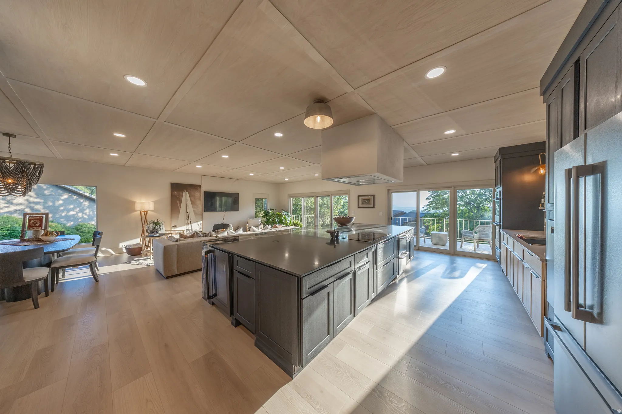 Open-concept kitchen and great room featuring a large center island with dark cabinetry and stone countertop, stainless steel appliances, white cabinetry, wood-finish flooring, recessed ceiling lighting, and expansive glass doors and windows overlooking o
