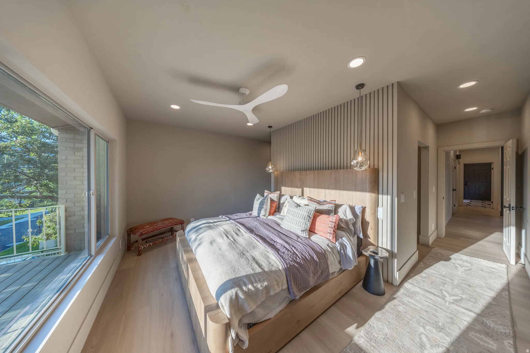 Contemporary primary bedroom showcasing a wood-finish accent wall with integrated pendant lighting, expansive windows with natural light, ceiling fan, recessed lighting, and wood-finish flooring throughout