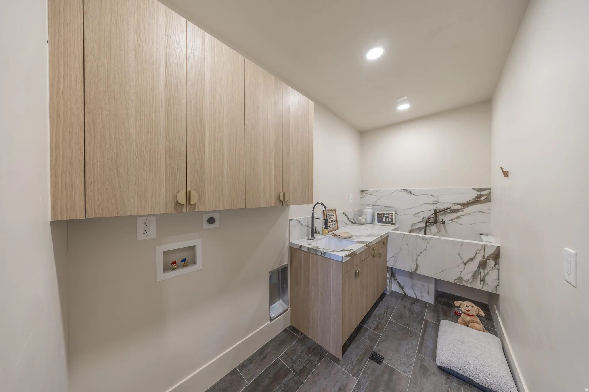 Modern laundry room featuring custom wood-finish cabinetry with ample storage, dual stainless steel sink with gooseneck faucet, dark tile flooring, and recessed lighting throughout