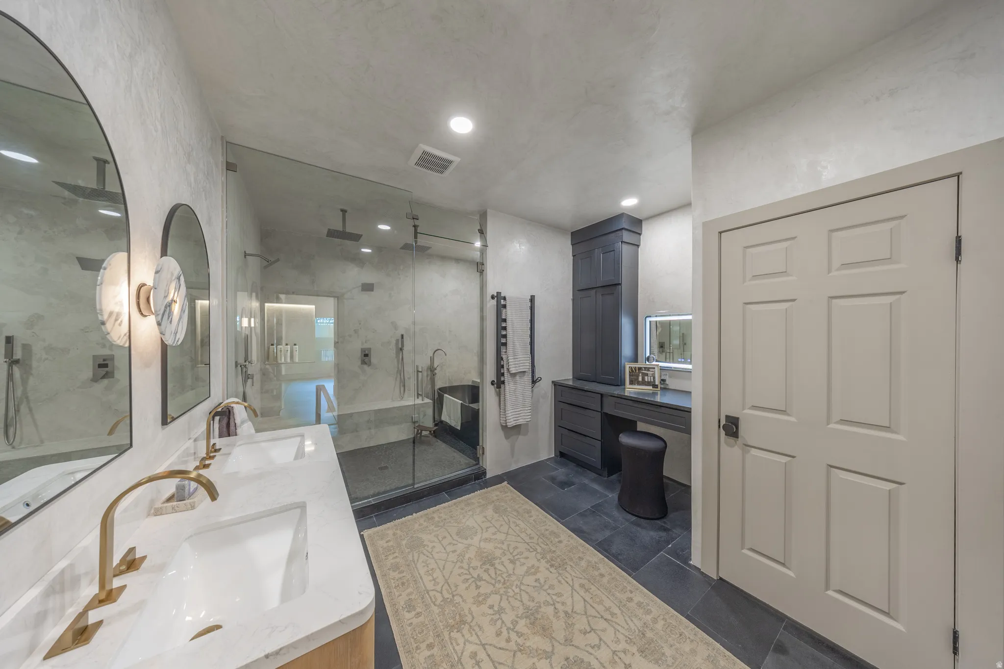 Primary Bathroom featuring a floating vanity with rectangular vessel sink, quartz countertop, and brass faucet; large round illuminated mirror; walk-in shower with frameless glass enclosure and tile surround; dual vanity layout with ample countertop stora