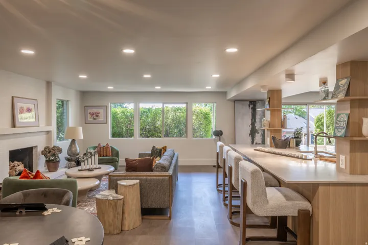 Recessed lighting throughout, a fireplace with a light-colored brick surround, wood-finish flooring, multiple windows providing natural light, and a built-in bar with open shelving
