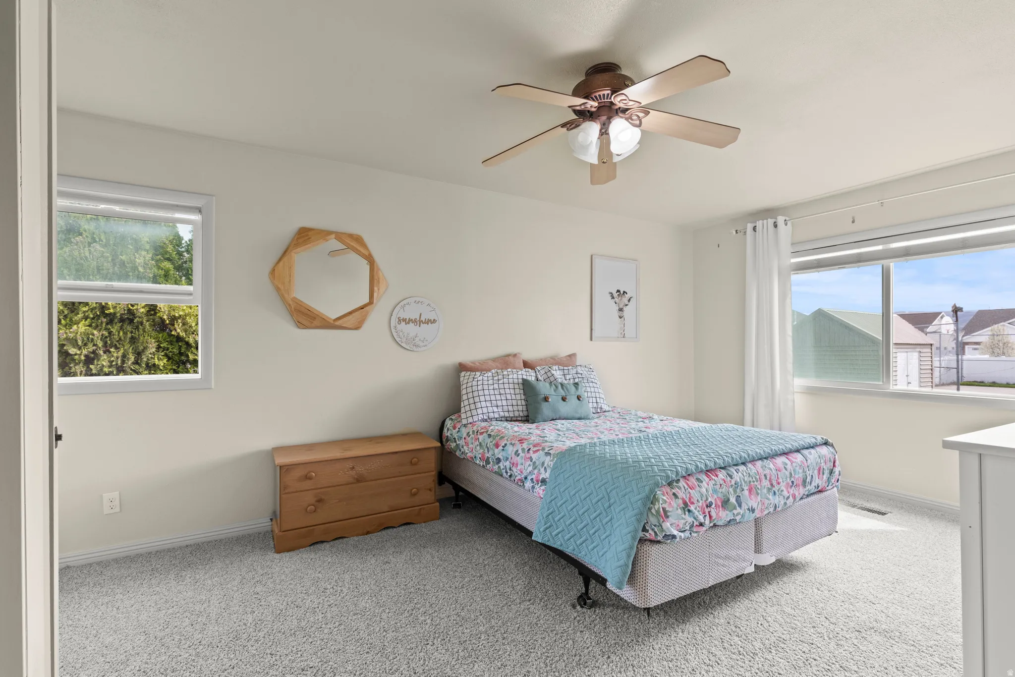 Bright secondary bedroom featuring carpet flooring, ceiling fan with integrated lighting, wood-finish nightstand, and dual windows with natural light and exterior views