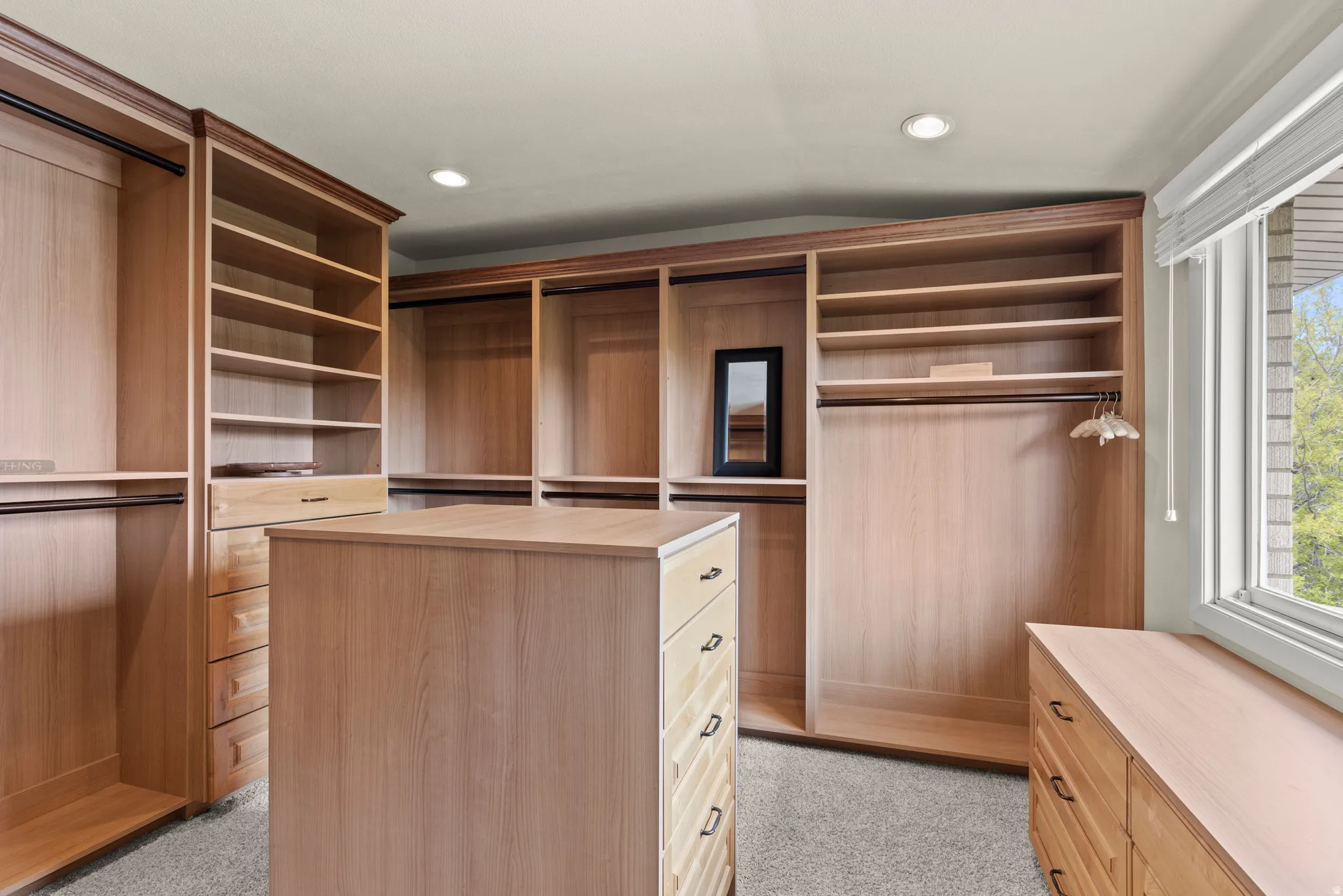 Luxurious walk-in closet featuring rich wood cabinetry throughout, custom shelving with varied depths for optimal storage, a central island dresser with multiple drawers, recessed lighting, and abundant natural light from a large window