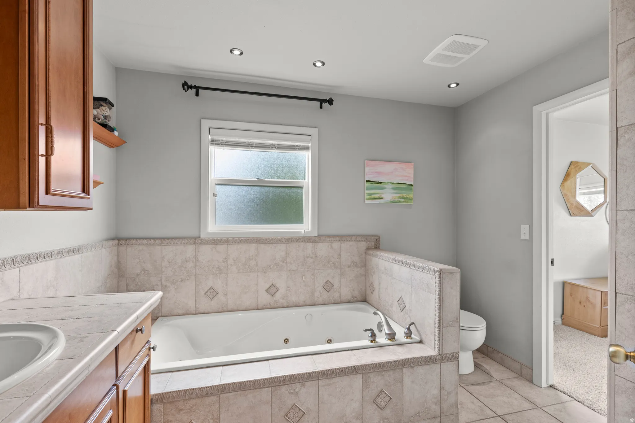 Spa-inspired primary bathroom featuring a deep soaking tub with tile surround, dual vanity with wood-finish cabinetry, recessed ceiling lighting, and a large window providing natural light