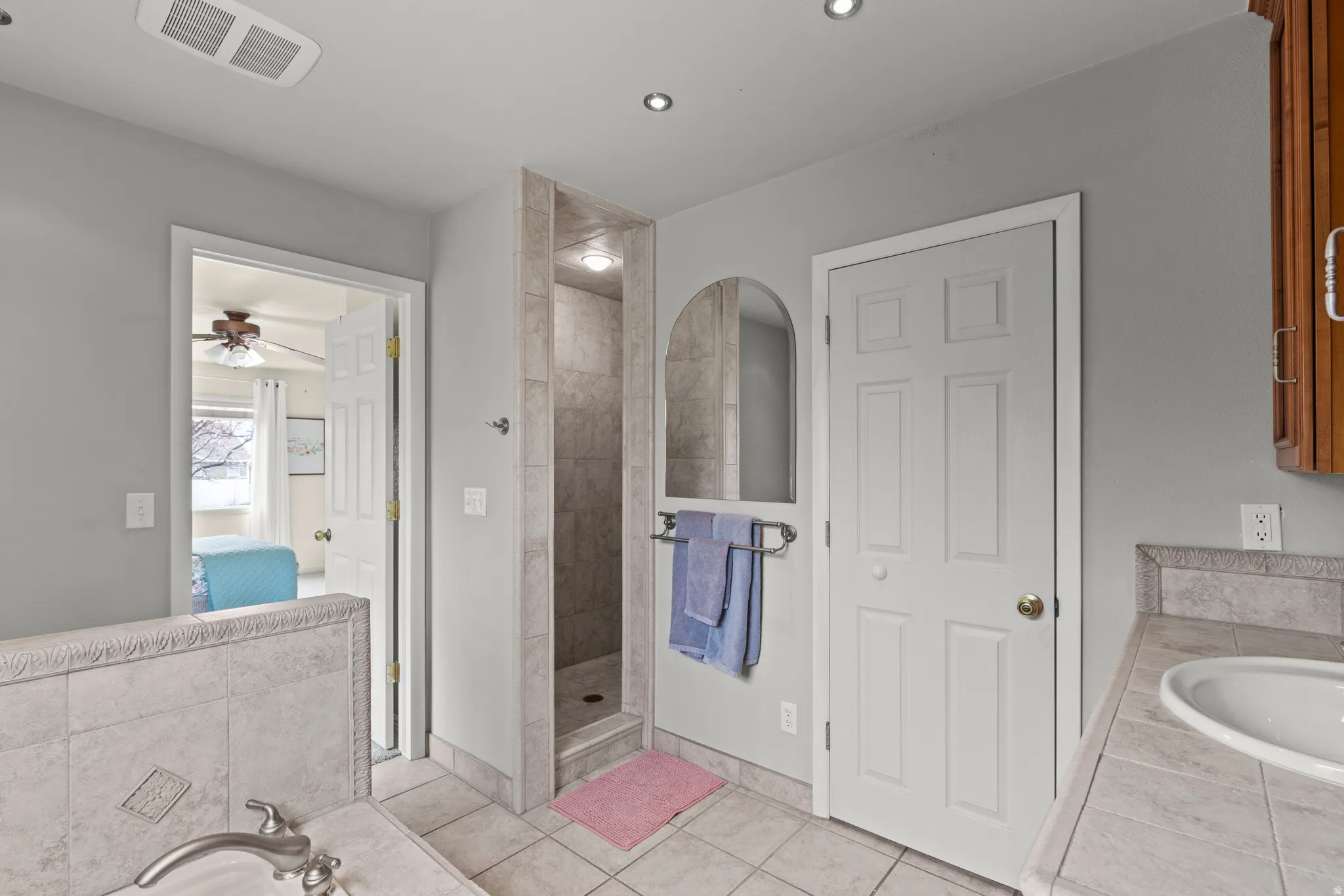 Spa-inspired primary bathroom featuring a walk-in shower with tile surround and rainfall showerhead, dual vanity with ceramic sink and integrated lighting, heated towel rack, recessed ceiling fixtures, and neutral tile flooring
