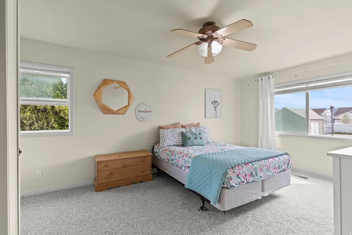 Bright secondary bedroom featuring carpet flooring, ceiling fan with integrated lighting, wood-finish nightstand, and dual windows with natural light and exterior views