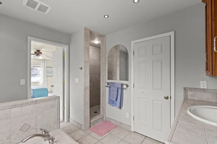 Spa-inspired primary bathroom featuring a walk-in shower with tile surround and rainfall showerhead, dual vanity with ceramic sink and integrated lighting, heated towel rack, recessed ceiling fixtures, and neutral tile flooring