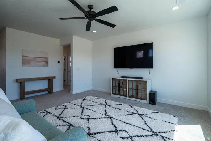 Spacious primary living area featuring a wall-mounted flat-screen television above built-in cabinetry with glass-front storage, ceiling fan with integrated lighting, geometric area rug, and abundant natural light from recessed ceiling fixtures
